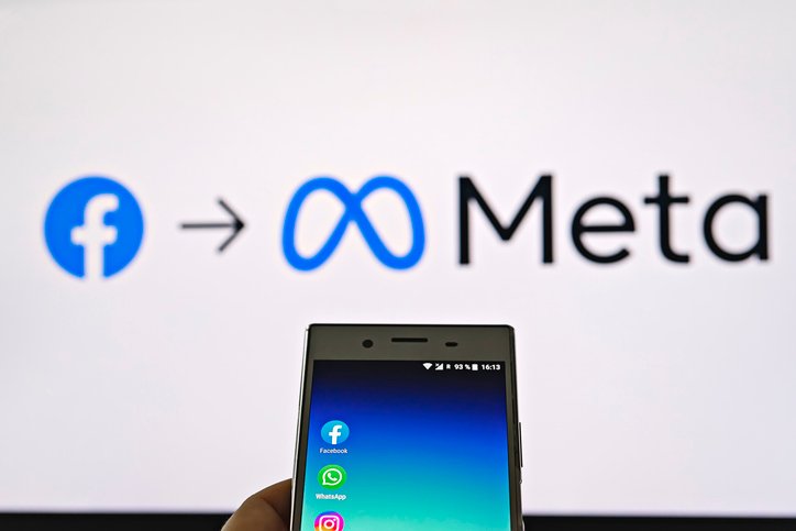 Mobile display with logo of Facebook, WhatsApp and Instagram apps in hand against blurred META logotype on white monitor